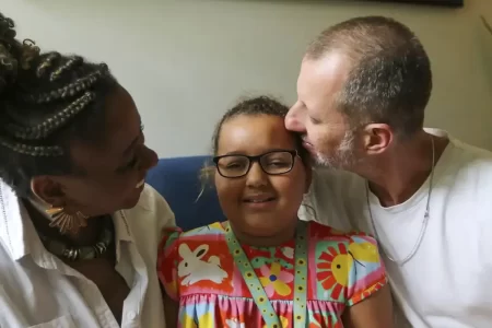 Flora sorrindo ao lado dos pais, mostrando força e alegria durante o tratamento