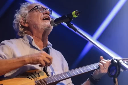 Renato Teixeira tocando violão durante apresentação ao vivo, com cenário que reflete a turnê Estrada Eu Sou.