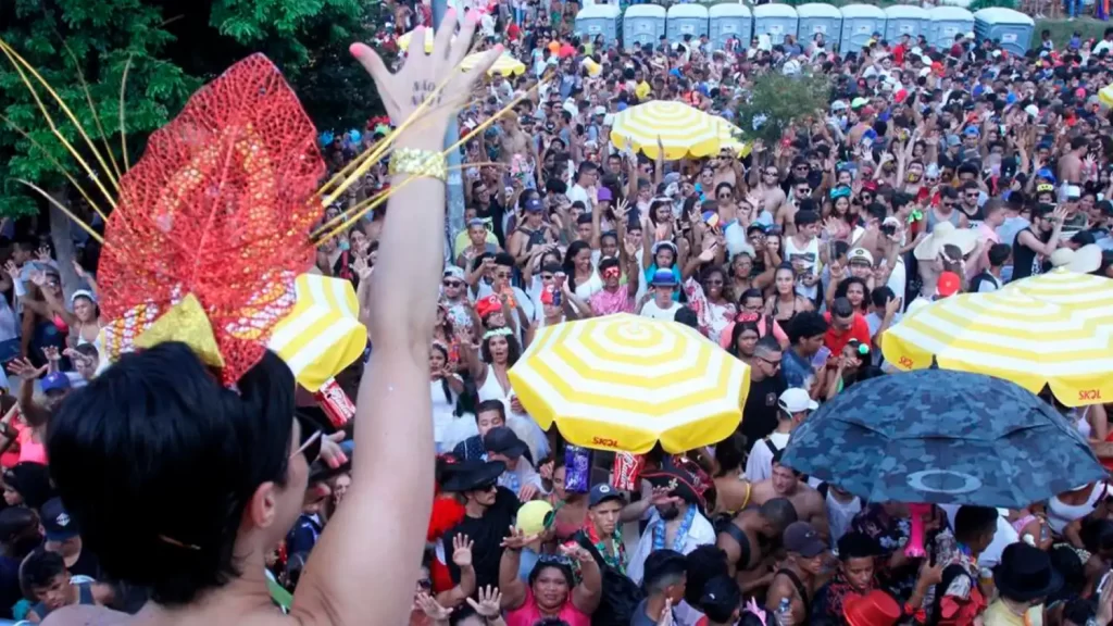 Pessoas se divertindo no Carnaval com máscaras e preservativos distribuídos em ação de conscientização da Secretaria de Saúde.