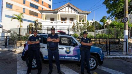 Três agentes da Polícia Municipal de São Bernardo do Campo posam em frente a uma viatura estacionada. Ao fundo, A Casa de Passagem para mulheres vítimas de violência, destacando a presença da GCM na proteção e segurança do local.