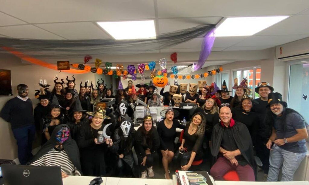 Equipe da MBigucci fantasiada no Halloween, com decoração temática, doces e cartazes do concurso interno.